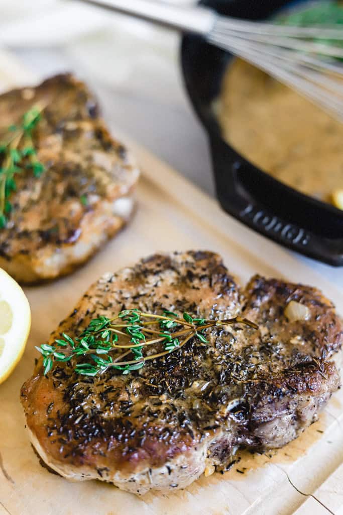 A close up shot of a pork chop on a cutting board topped with fresh thyme and a skillet of lemon garlic cream sauce behind it