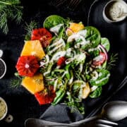 A bowl of greens topped with blood oranges and a poppyseed dressing on a black background.