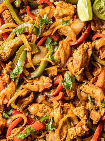 An overhead closeup shot of chicken fajitas on a pan topped with chopped cilantro and lime wedges.