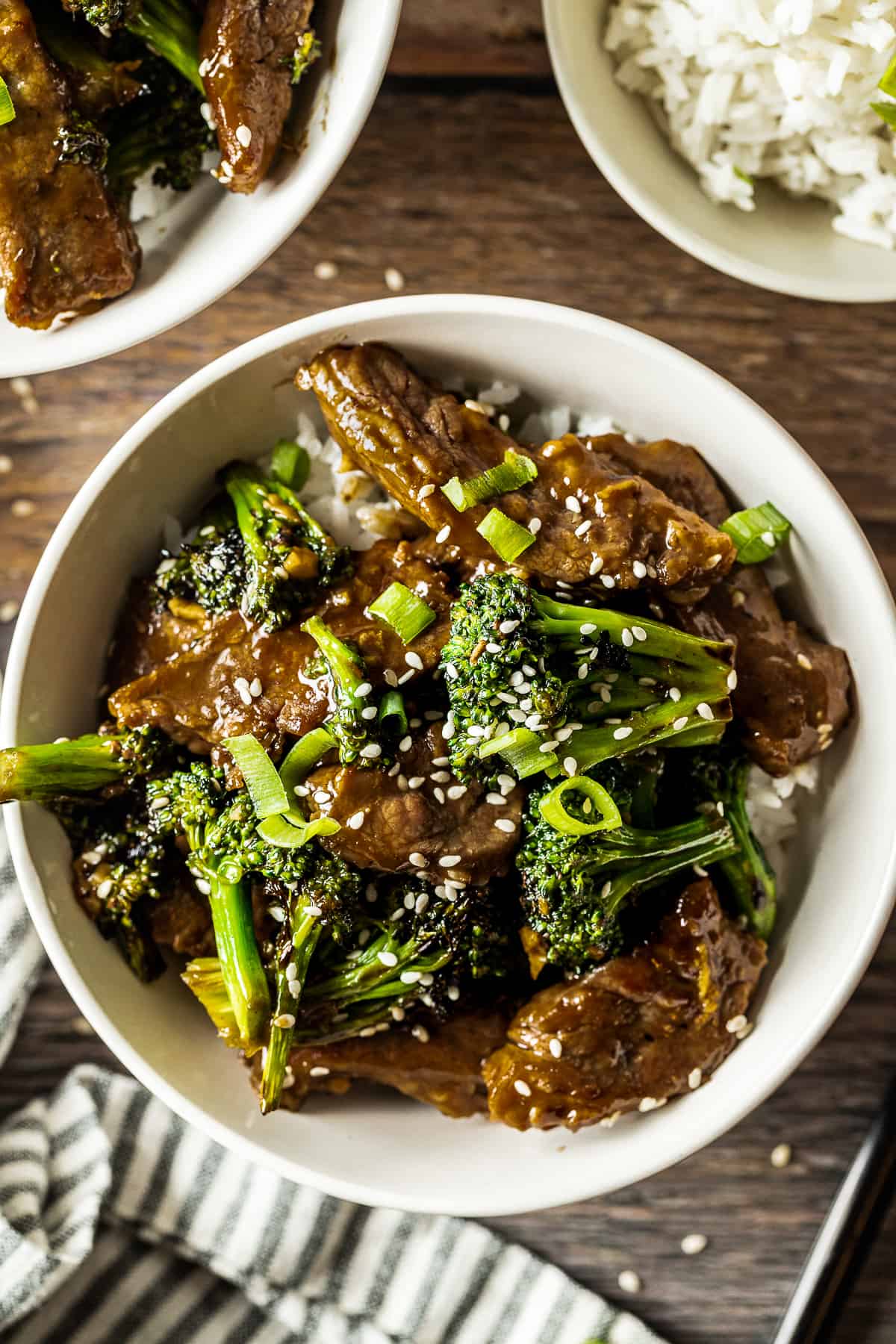 A white bowl of beef and broccoli on a wood background near a bowl of rice and a blue and white linen.