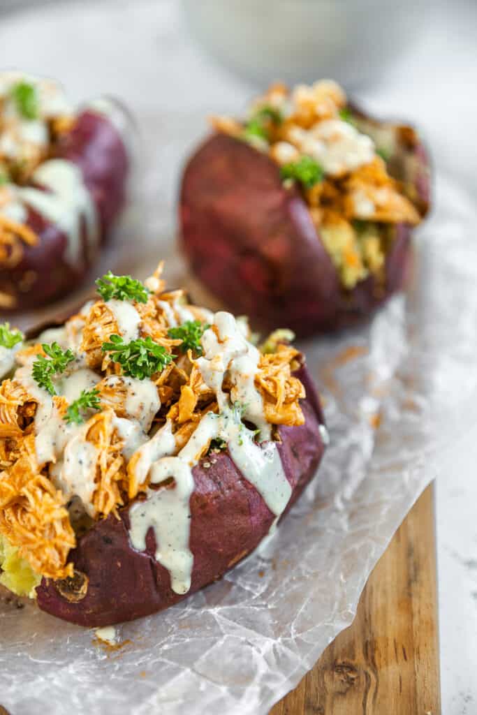 Three buffalo ranch chicken stuffed sweet potatoes on a cutting board topped with ranch and parsley