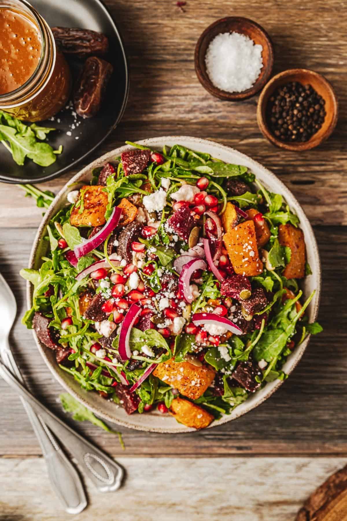 An overview shot of a bowl of a roasted butternut and beetroot salad on a wood background near utinsels and dressing
