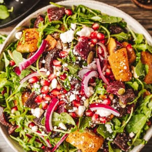 An overview shot of a bowl of a roasted butternut and beetroot salad on a wood background near utinsels and dressing