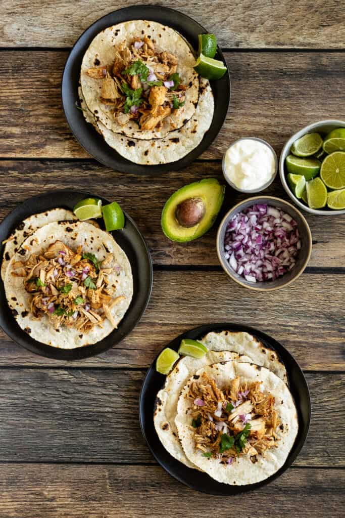An overview shot of three plates of tortillas topped with carnitas next to onions, avocado, and limes on a wood background