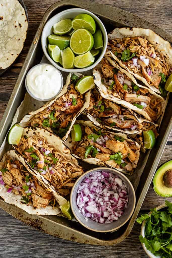 An overview shot of instant pot carnitas in tacos next to onions, limes, and sour cream on a wood backrgound