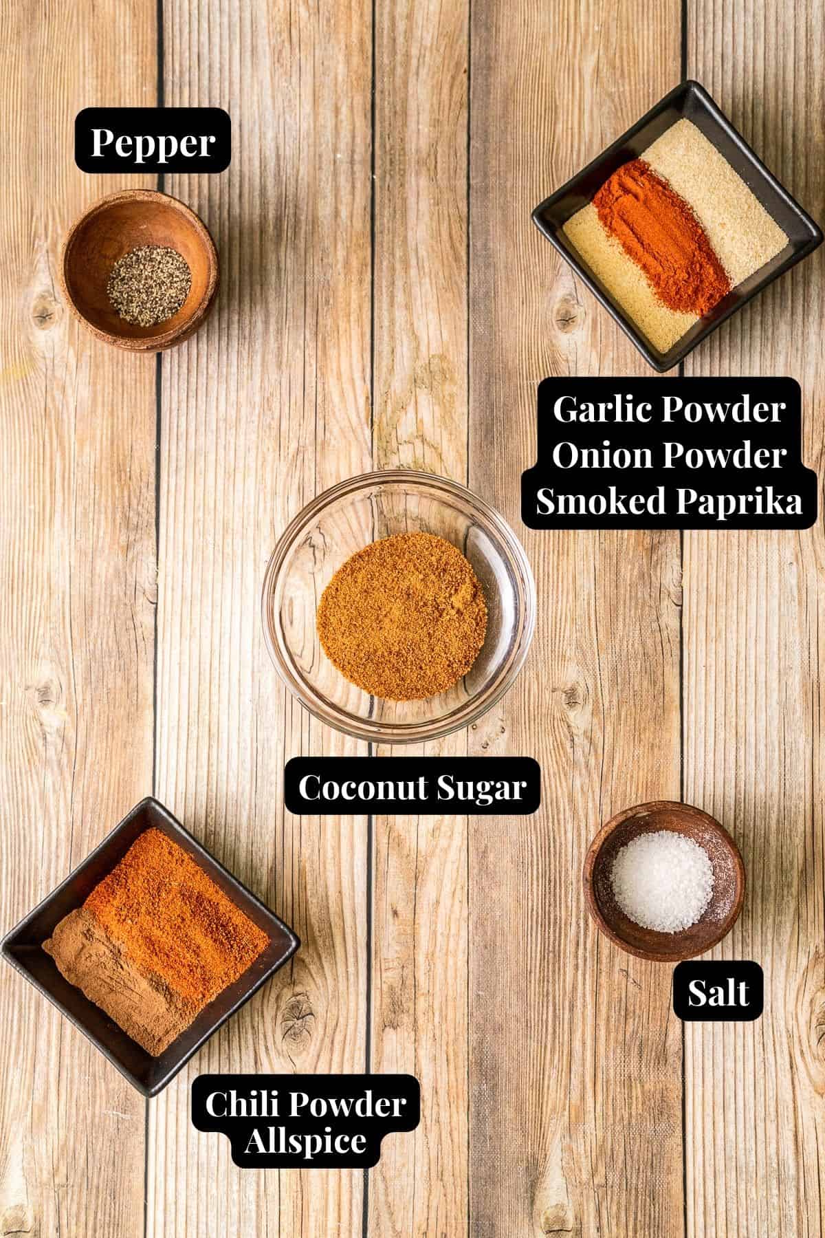 An overview shot of the spices and ingredients needed for chicken dry rub in various containers on a wood background.