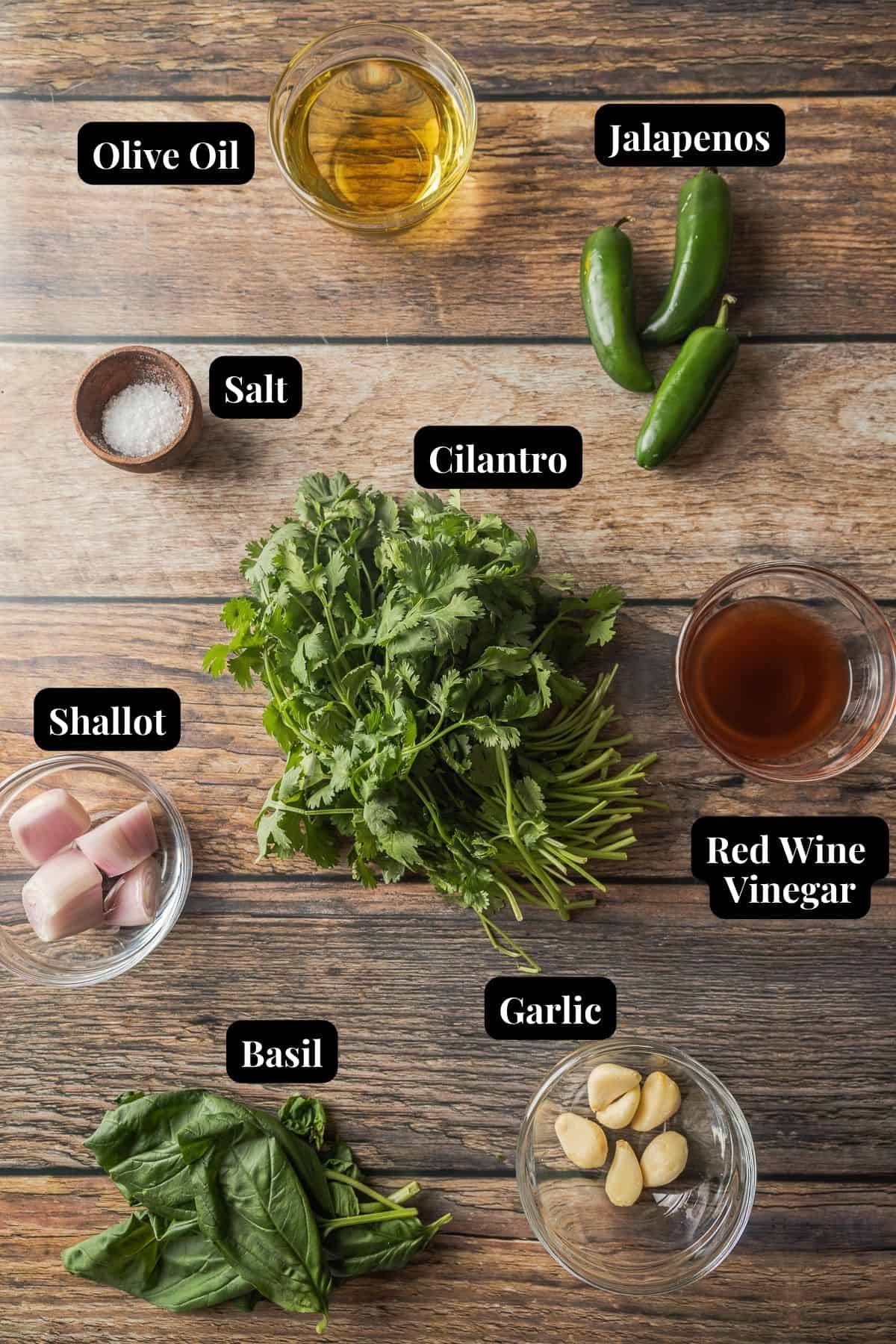 An overview shot of the ingredients needed for chimichurri with basil in glass bowls on a wood background.