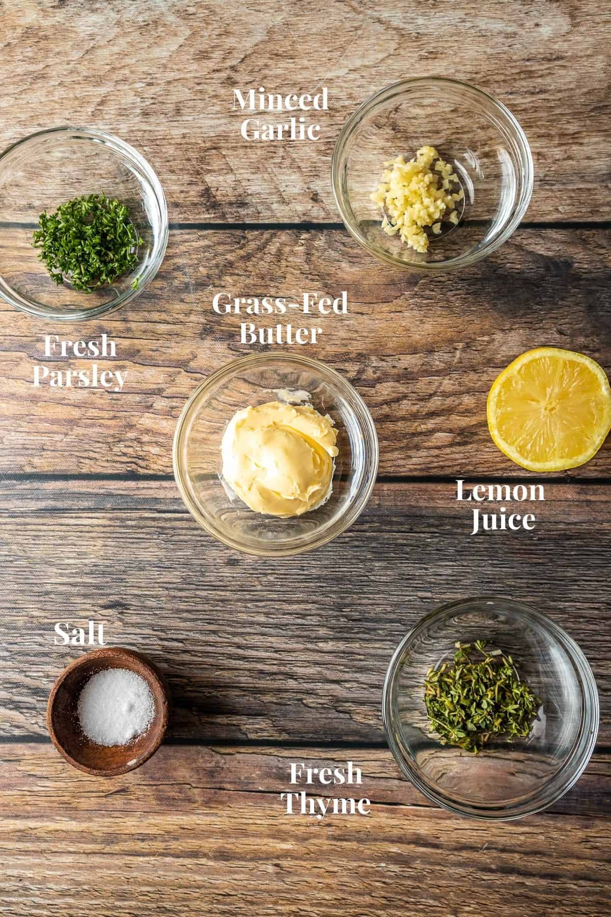 An overview shot of the ingredients needed for an herb and garlic butter compound on a wood background.