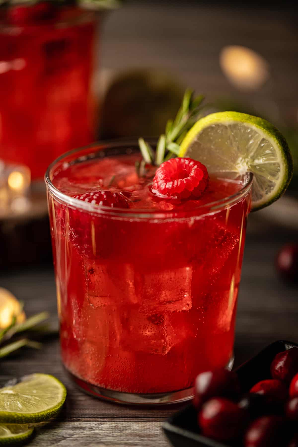 A glass filled with a cranberry raspberry mocktail topped with raspberries, rosemary, and limes on a wood background