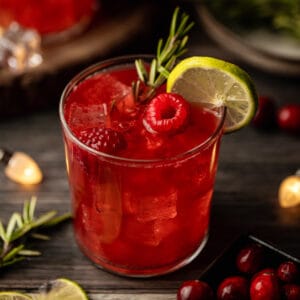 A glass filled with a cranberry raspberry mocktail topped with raspberries, rosemary, and limes on a wood background