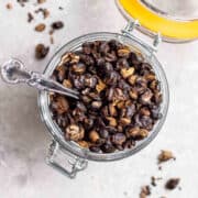 A glass jar of crispy black beans with a silver spoon on a grey background.