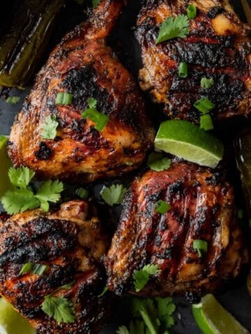 Jerk chicken thighs topped with cilantro and lime wedges on a black cutting board.