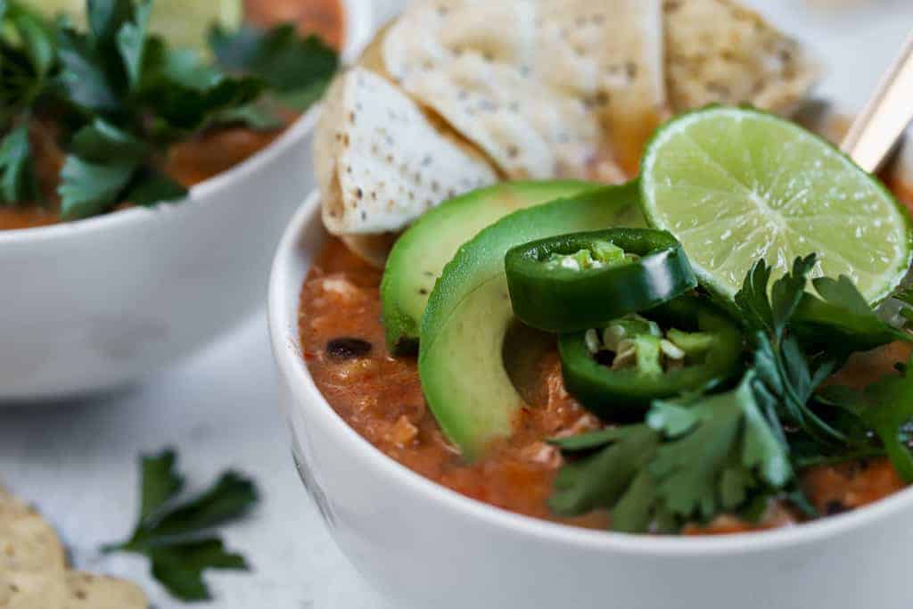 Two bowls of chicken tortilla soup topped with chips, limes, and avocado