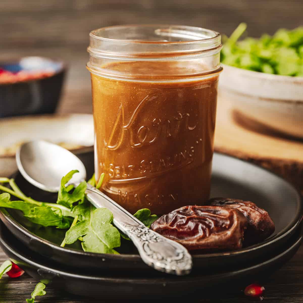 A mason jar of Whole30 date and dijon dressing on a black plate near greens