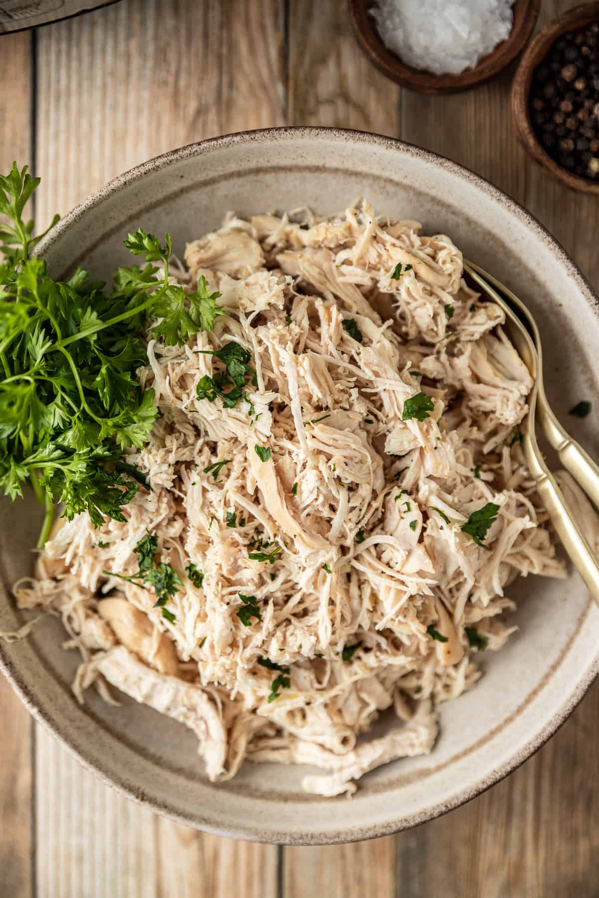 An overview shot of a bowl of plain shredded chicken topped with chopped parsley in a beige bowl.