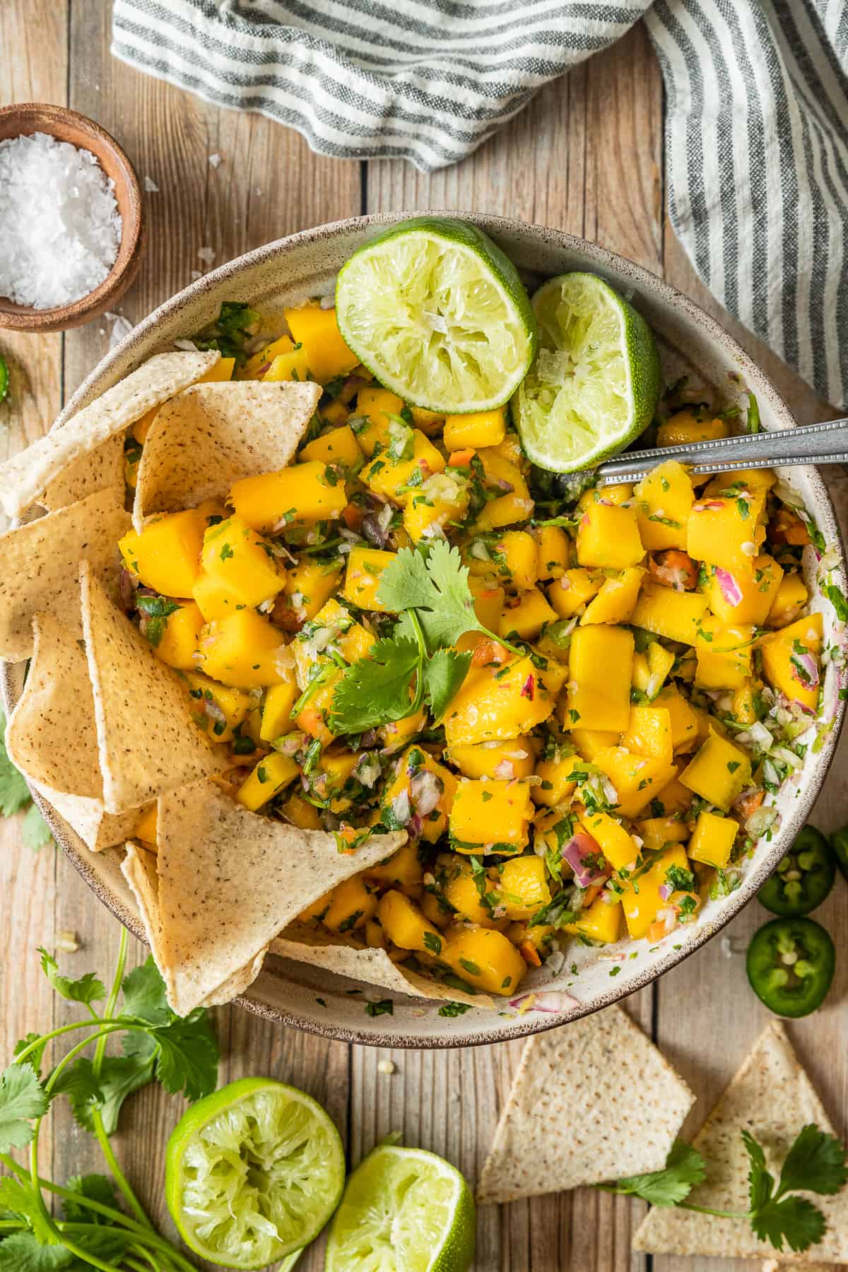 A bowl of mango salsa on a wood background topped with lime and cilantro near a bowl of chips.
