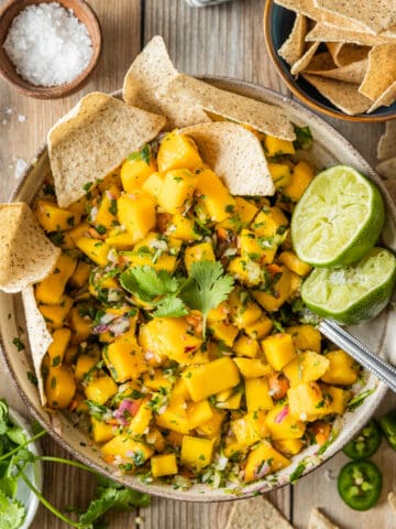 A top down shot of a bowl of mango salsa topped with cilantro, lime, and served with chips on a wood background.