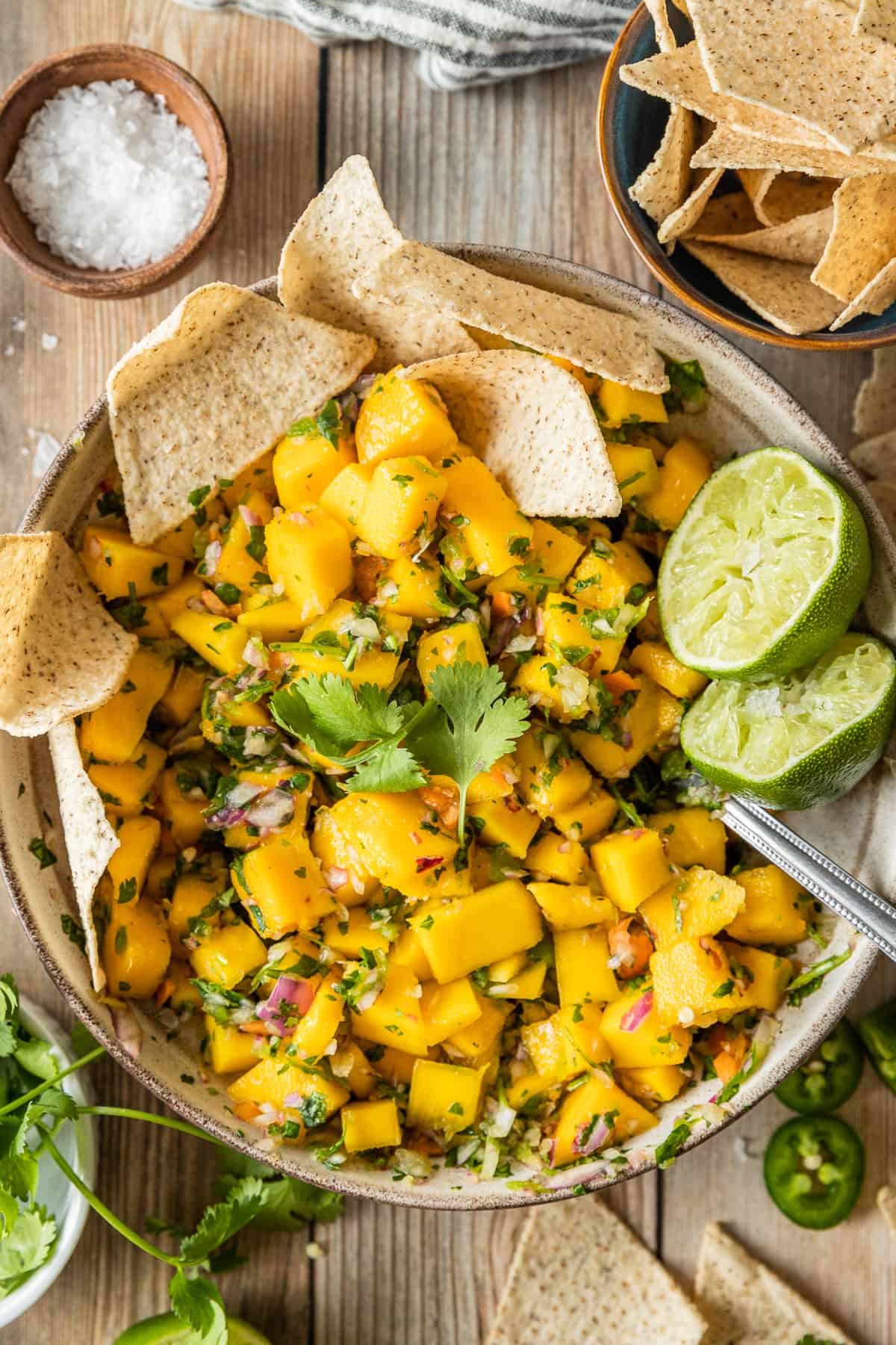 A top down shot of a bowl of mango salsa topped with cilantro, lime, and served with chips on a wood background.  