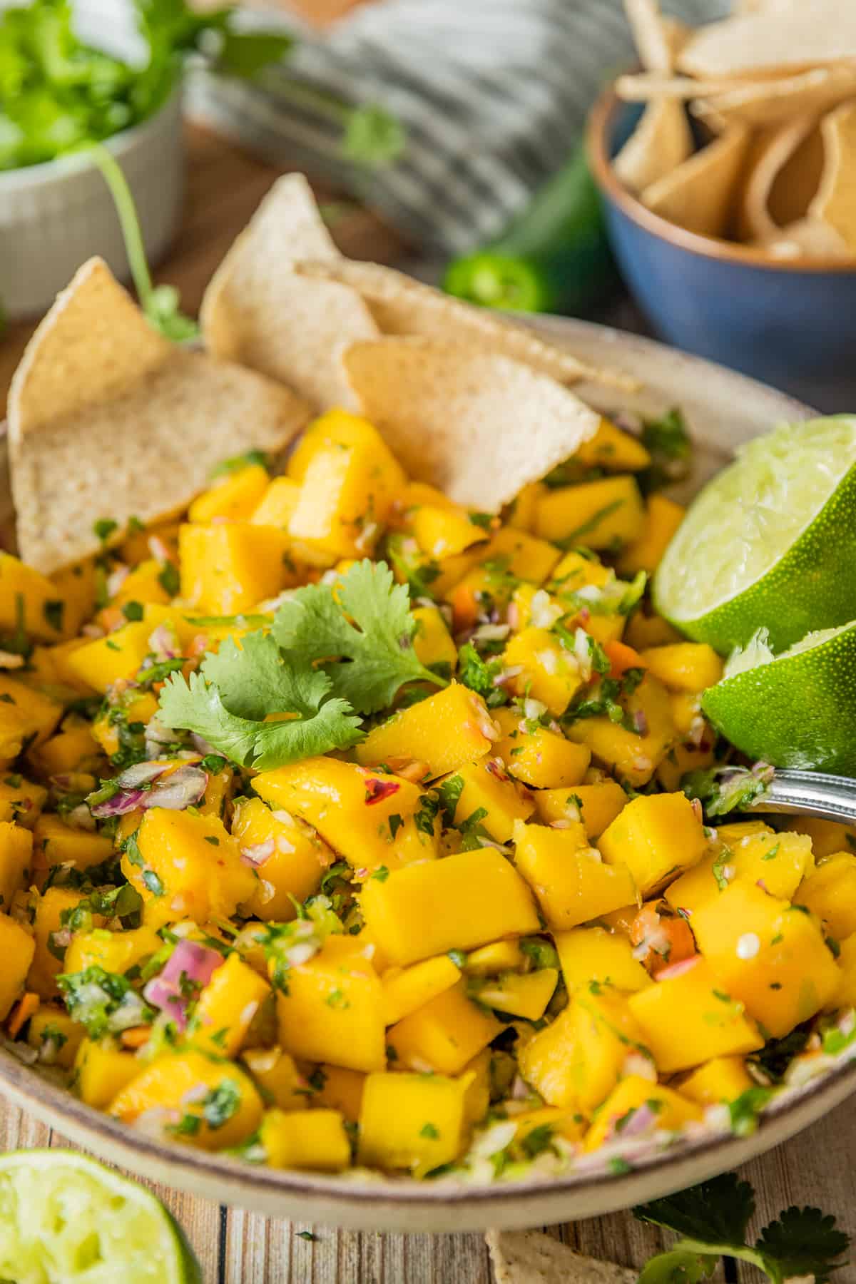 A bowl of mango salsa on a wood background topped with lime and cilantro near a bowl of chips.