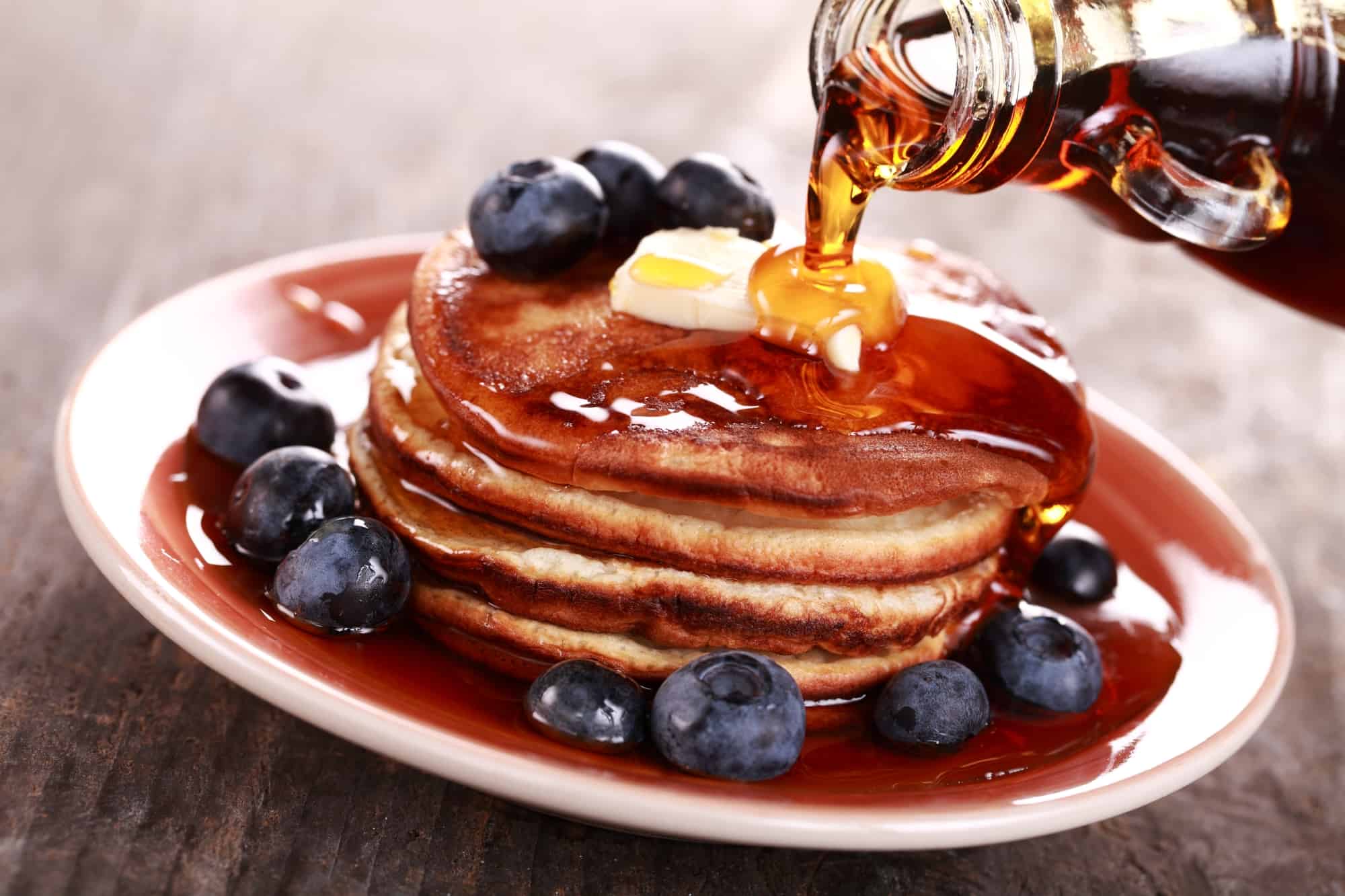 A jar of maple syrup being drizzled onto a stack of pancakes topped with blueberries on a nuetral colored background.