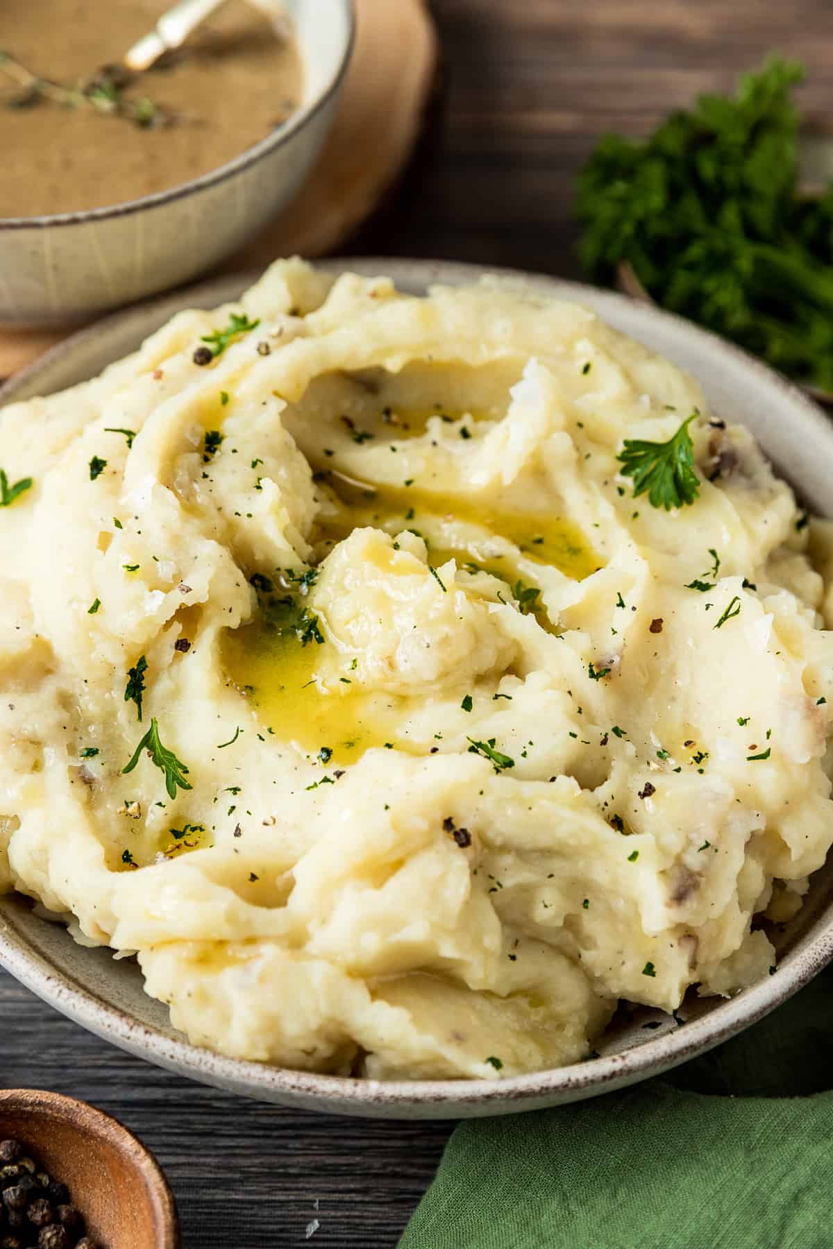 A 45 degree shot of a bowl of mashed potatoes topped with melted butter, pepper, and parsley on a wood backrgound next to a bowl of gravy and pepper.