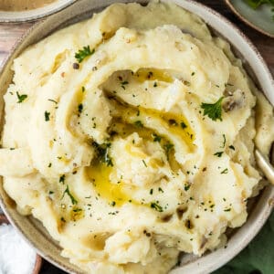 An overview shot of a bowl of mashed potatoes topped with melted butter, pepper, and parsley on a wood backrgound next to a bowl of gravy and pepper.