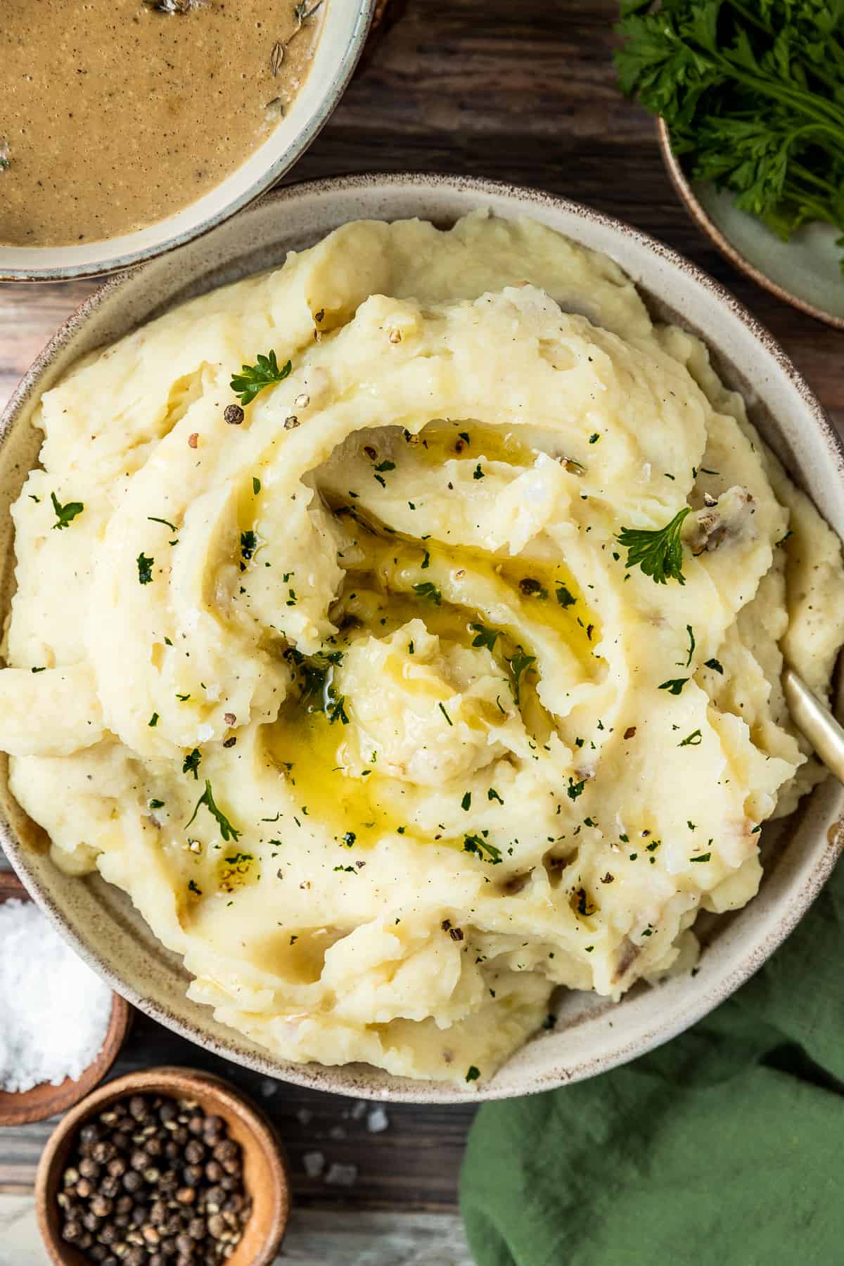 An overview shot of a bowl of mashed potatoes topped with melted butter, pepper, and parsley on a wood backrgound next to a bowl of gravy and pepper.
