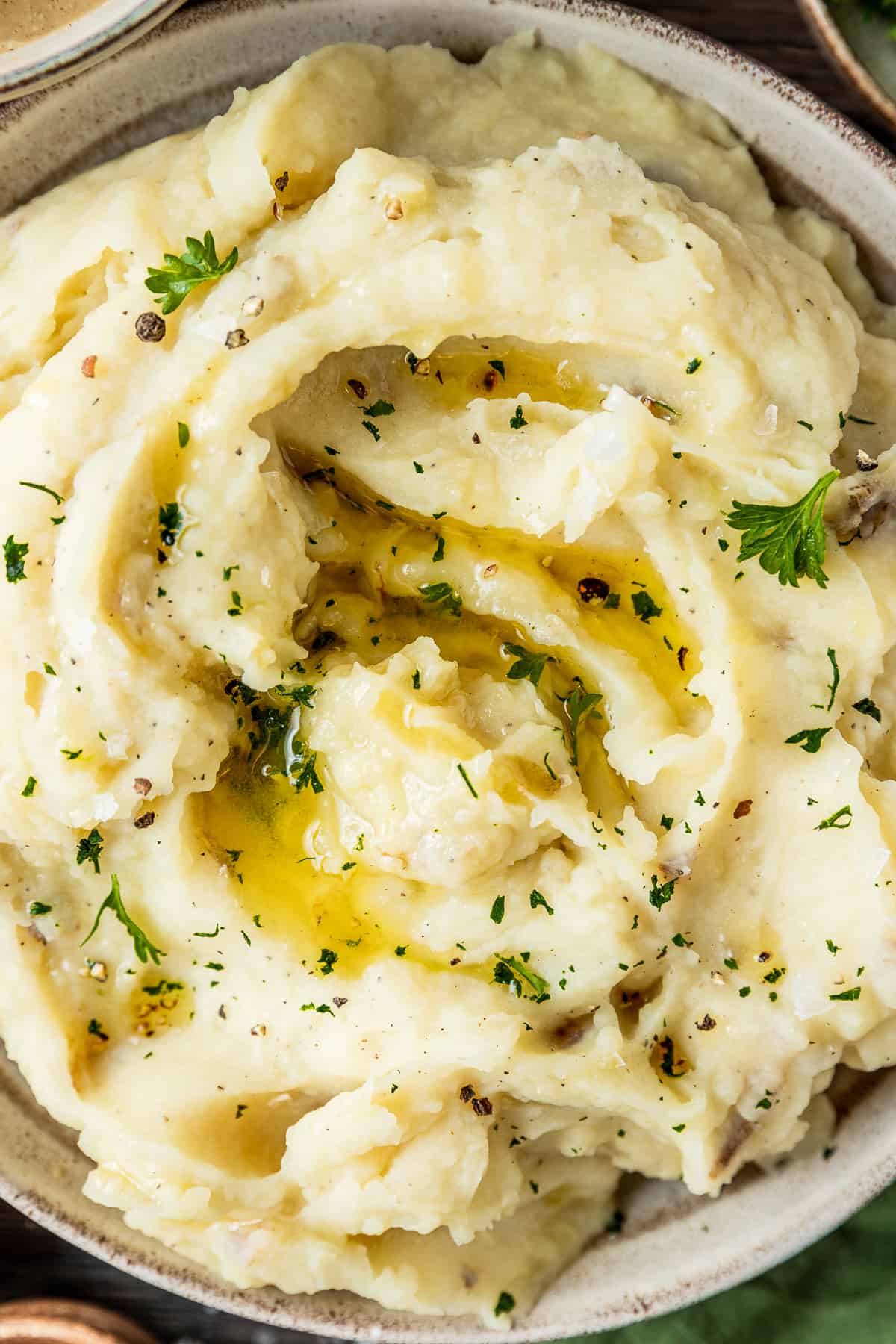 An overview shot of a bowl of mashed potatoes topped with melted butter, pepper, and parsley on a wood backrgound next to a bowl of gravy and pepper.