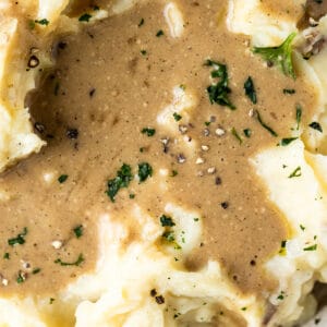 A bowl of mashed sweet potatoes with a pool of gravy and topped with parsley, salt and pepper on a wood background.