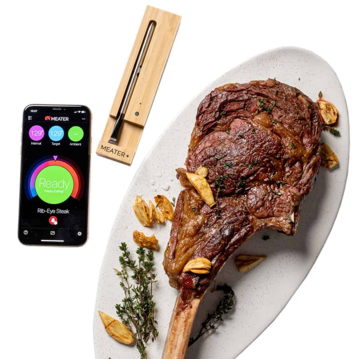 A meater+ thermometer near a phone showing the app and a piece of steak on a white background.