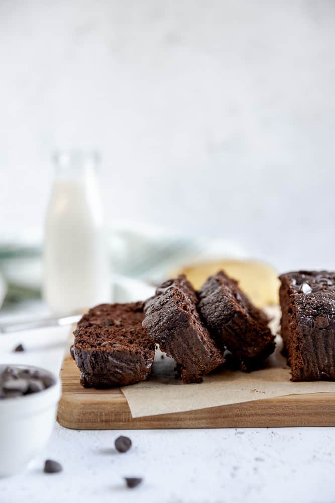 A cutting board with chocolate chip banana bread slices in front of a glass of milk and grey background