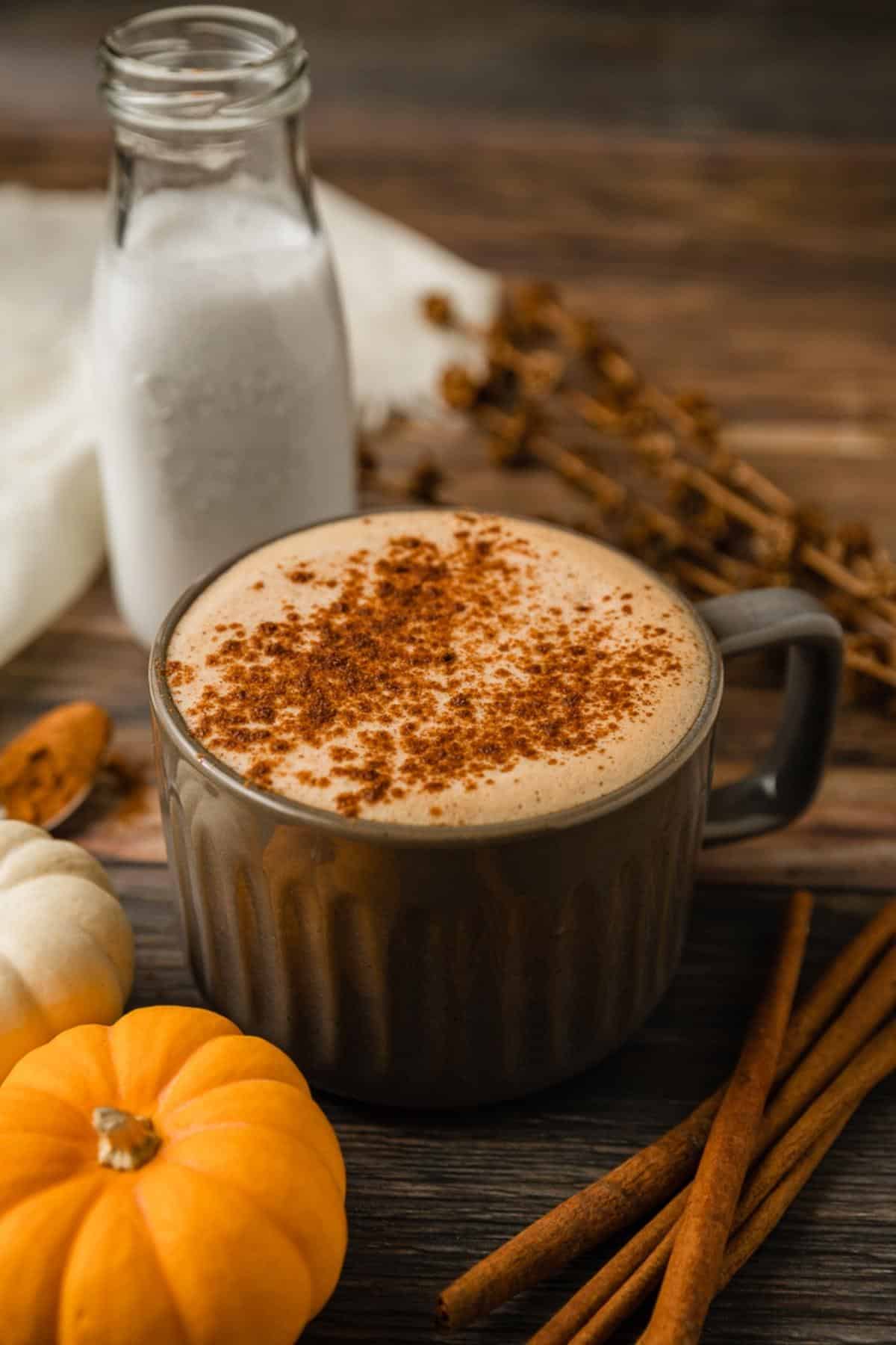 A grey mug filled with a paleo pumpkin spice latte on a wood background next to cinnamon sticks and pumpkins.