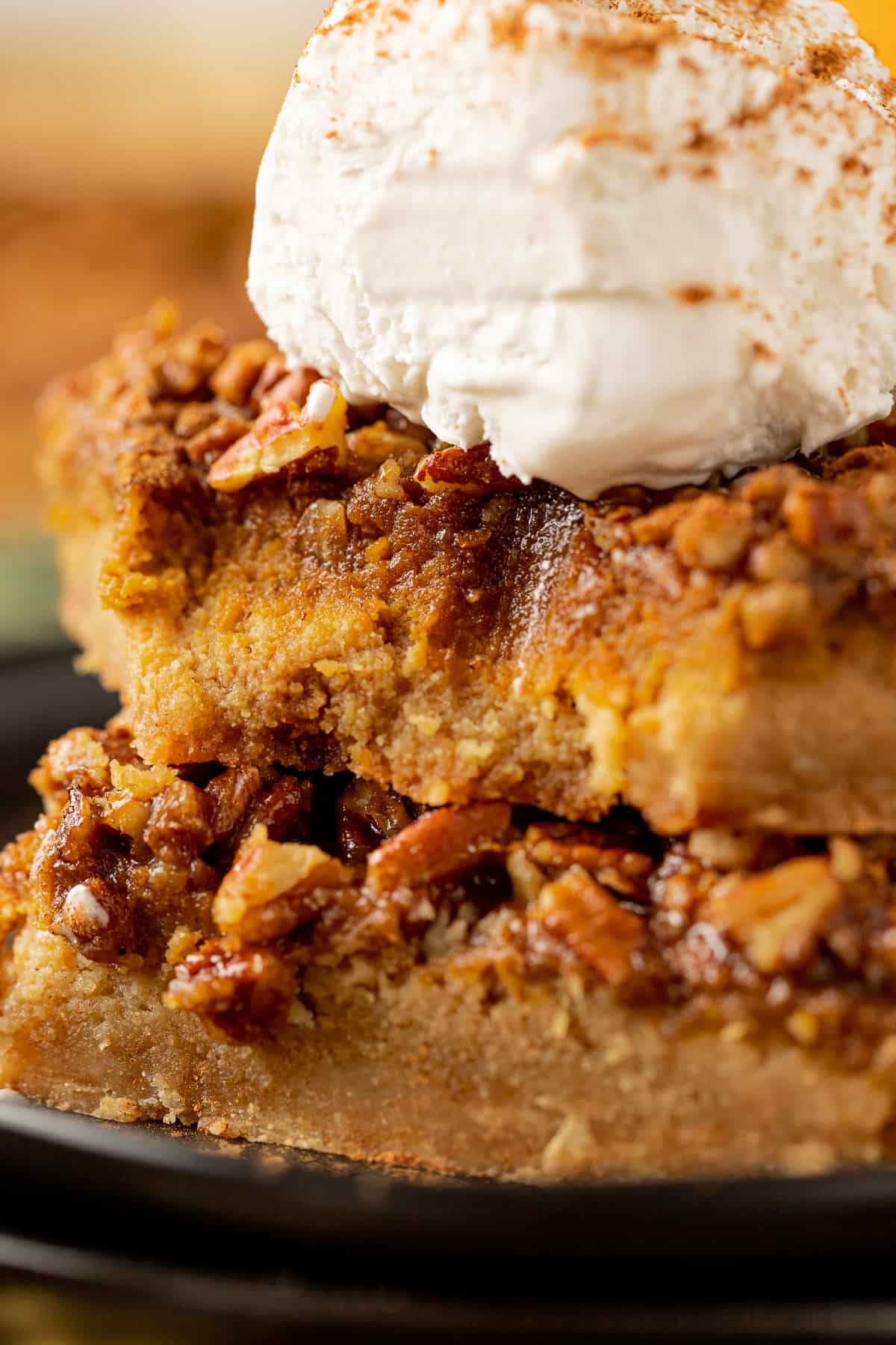 A stack of pumpkin pecan pie bars on a black plate with a bite removed topped with whipped cream and pumpkin pie spice