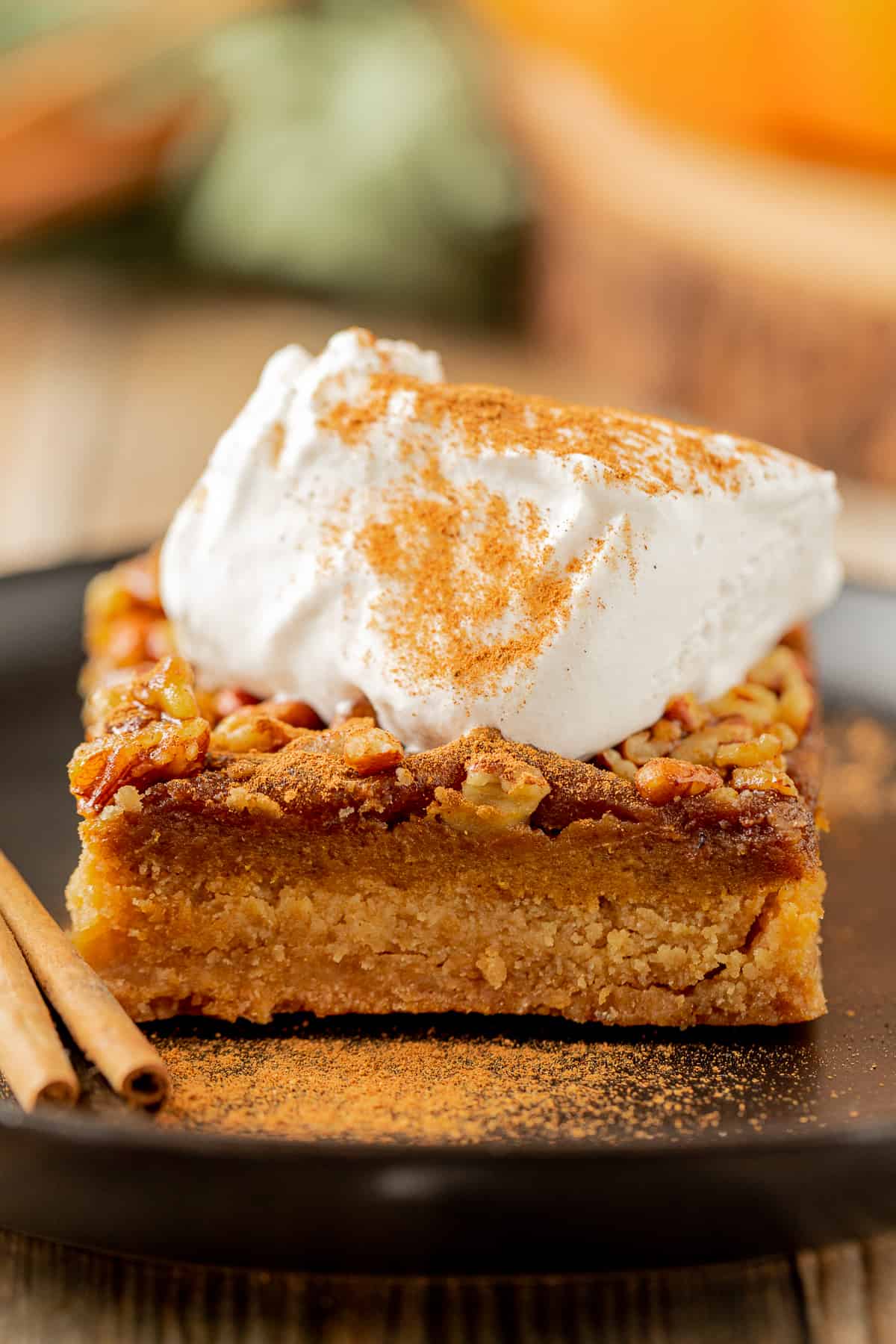 A head on view of a pumpkin pie bar with pecan pie topping, topping with coconut whipped cream, showing the three layers of the bar on a black plate