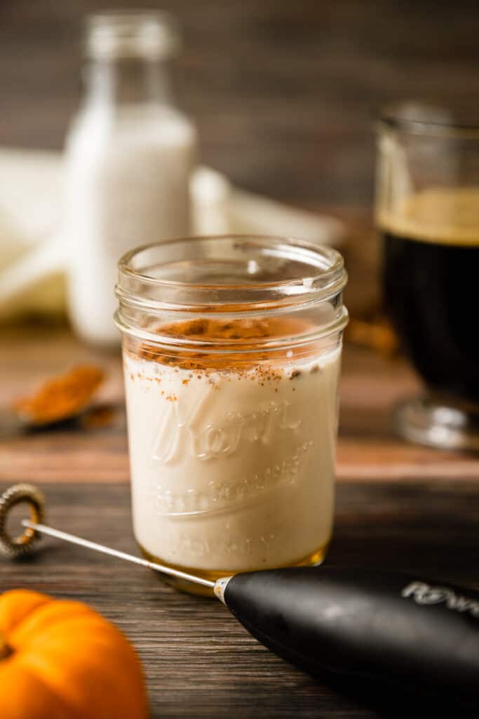 A jar of steamed milk and pumpkin spice near a frother on a wood background