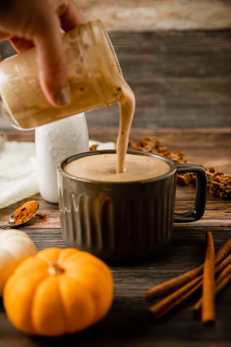 A jar of frothy steamed milk being poured into a mug of coffee on a wood background