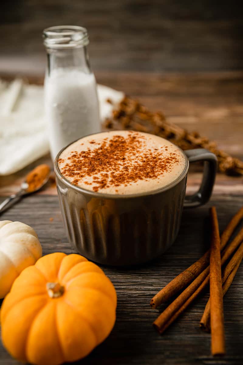 A grey mug filled with a paleo pumpkin spice latte on a wood background next to cinnamon sticks and pumpkins.