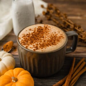 A grey mug filled with a paleo pumpkin spice latte on a wood background next to cinnamon sticks and pumpkins.