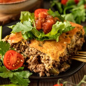 A square piece of taco casserole topped with tomatoes, avocado, and lettuce on a black plate.