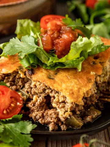 A square piece of taco casserole topped with tomatoes, avocado, and lettuce on a black plate.