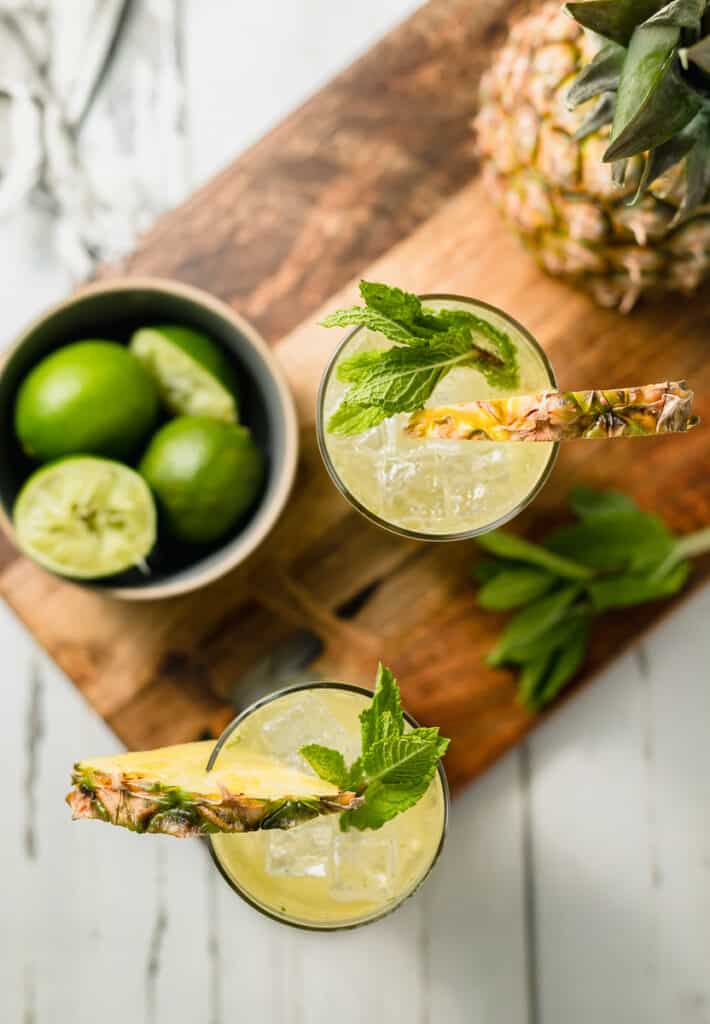 An overview shot of two mojito mocktails on a cutting board next to a pineapple and bowl of limes