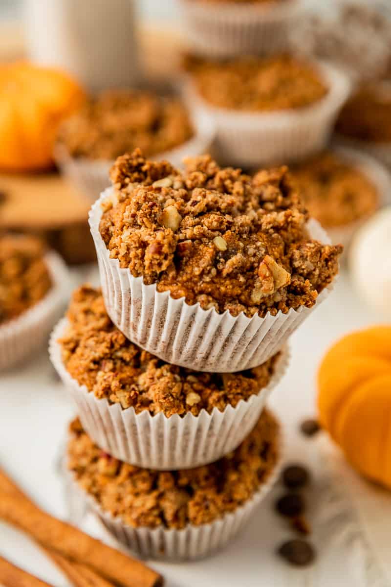 A stack of paleo pumpkin chocolate chip muffins on a white wood background near cinnamon sticks and chocolate chips