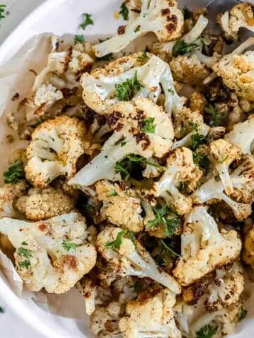 A bowl of roasted cauliflower on a cutting board next to a bunch of parsley.
