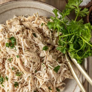 An overview shot of a bowl of plain shredded chicken topped with chopped parsley in a beige bowl