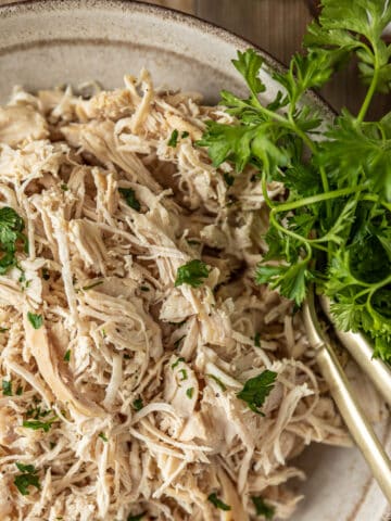 An overview shot of a bowl of plain shredded chicken topped with chopped parsley in a beige bowl