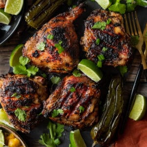 Jerk chicken thighs topped with cilantro and lime wedges on a black cutting board.