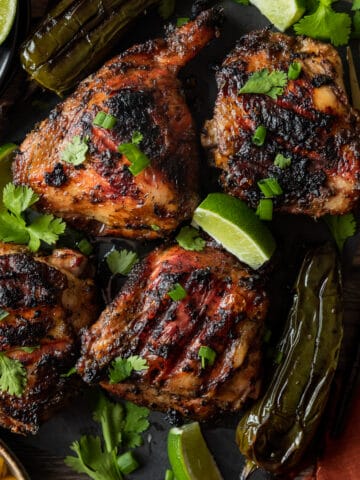 Jerk chicken thighs topped with cilantro and lime wedges on a black cutting board.