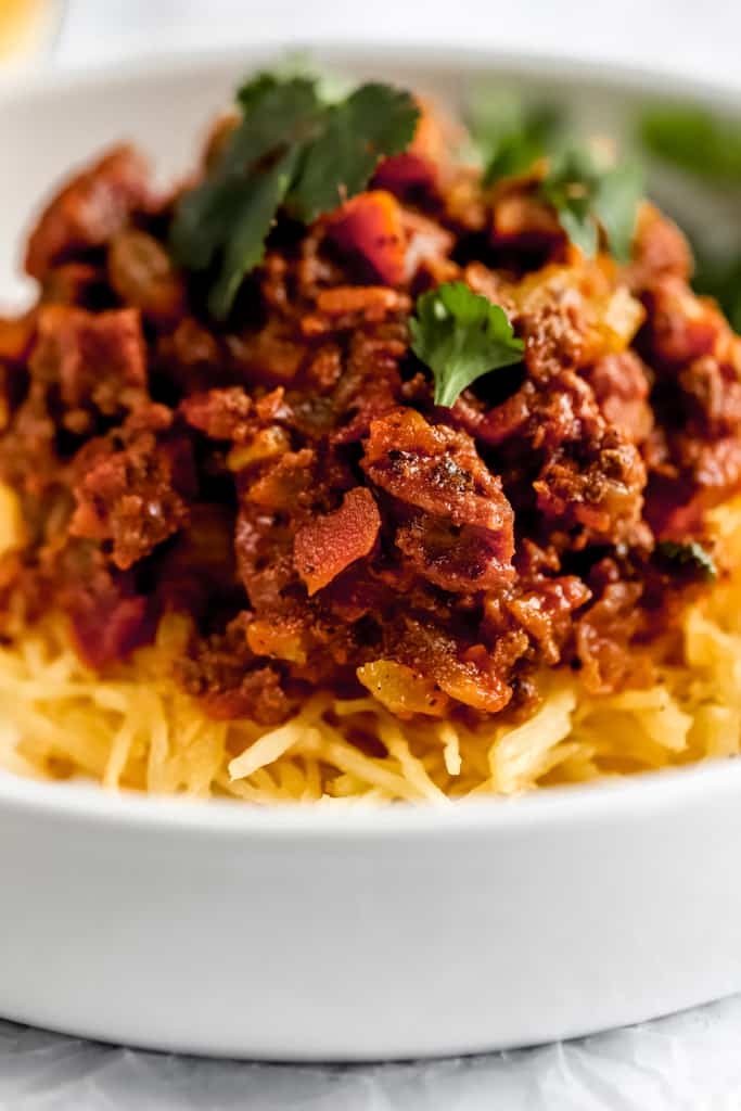 A close up view of a white bowl of al dente spaghetti squash topped with spaghetti meat sauce and chopped parsley.