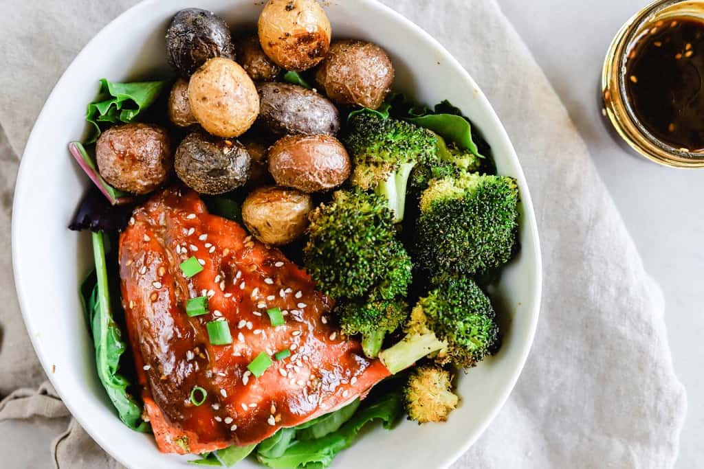 A bowl of teriyaki salmon with roasted potatoes and brocolli next to a jar of whole30 teriyaki sauce.