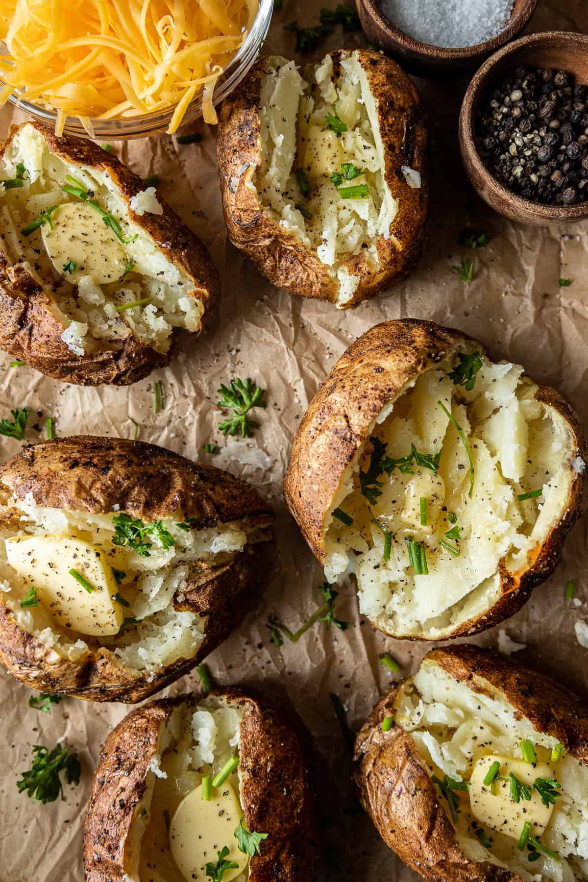 An overview shot of six smoked baked potatoes filled with melted butter, chives, and parsley next to a bowl of shredded cheese.